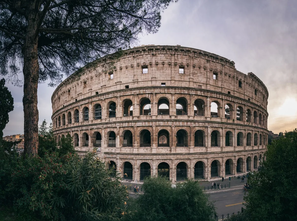 The Colosseum in Rome beneath a soft evening sky, framed by pine trees