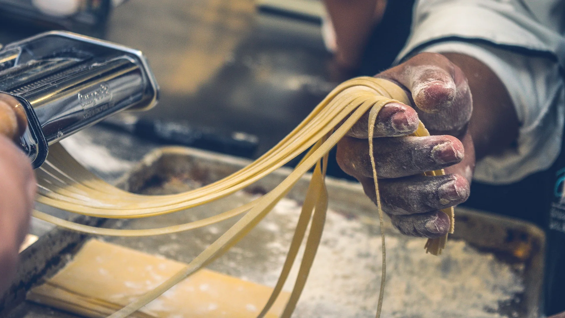 A flour-dusted hand lifts ribbons of fresh yellow tagliatelle beside a steel pasta machine on a floured tray